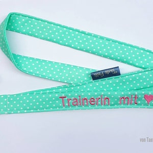 Puede incluir: Un cordón verde con lunares blancos y el texto "Trainerin mit" bordado en rosa con un corazón rosa. El cordón tiene una etiqueta azul que dice "Tante Pauli" y está unido a un silbato plateado.