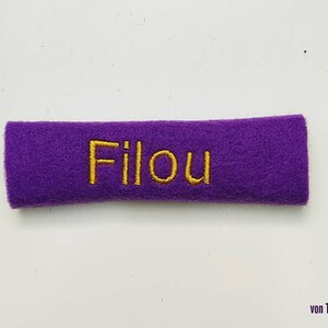 May include: A rectangular, purple felt patch with the embroidered word "Filou" in gold thread. The patch is a solid color and appears to be handmade. The text is centered on the patch.