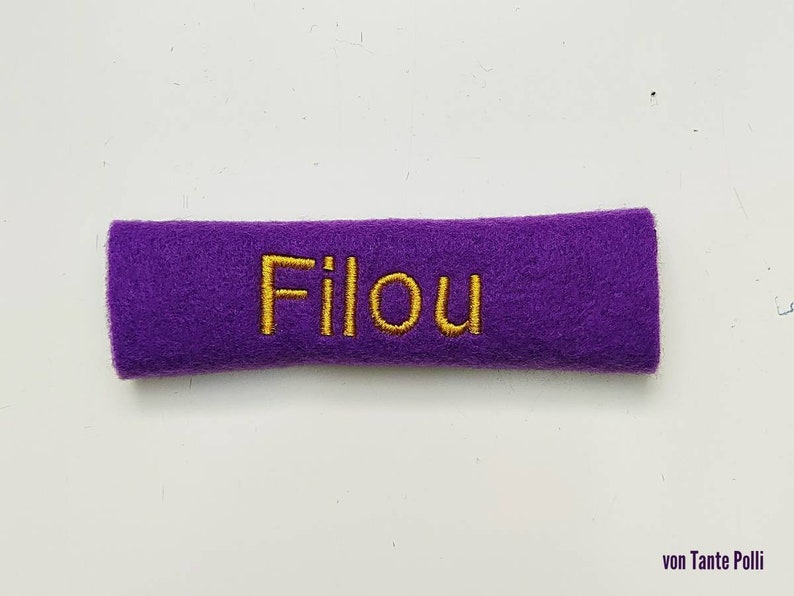 May include: A rectangular, purple felt patch with the embroidered word "Filou" in gold thread. The patch is a solid color and appears to be handmade. The text is centered on the patch.