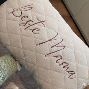 May include: Cream-colored, quilted pillow with the embroidered text "Beste Mama" in a cursive script. The pillow has a diamond pattern and a soft, textured appearance. The overall aesthetic is cozy and comforting.