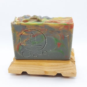 Puede incluir: Una pastilla de jab&oacute;n artesanal con un remolino de colores verde, amarillo, naranja y rojo. El jab&oacute;n tiene un dise&ntilde;o de faro en relieve en la parte delantera. El jab&oacute;n est&aacute; sentado en un jabonera de madera.