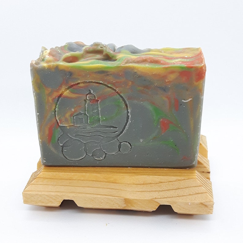 Puede incluir: Una pastilla de jab&oacute;n artesanal con un remolino de colores verde, amarillo, naranja y rojo. El jab&oacute;n tiene un dise&ntilde;o de faro en relieve en la parte delantera. El jab&oacute;n est&aacute; sentado en un jabonera de madera.