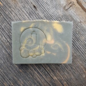 Puede incluir: Una pastilla de jab&oacute;n verde y amarilla con un logotipo de faro en relieve en la parte delantera. El jab&oacute;n est&aacute; sobre una superficie de madera.