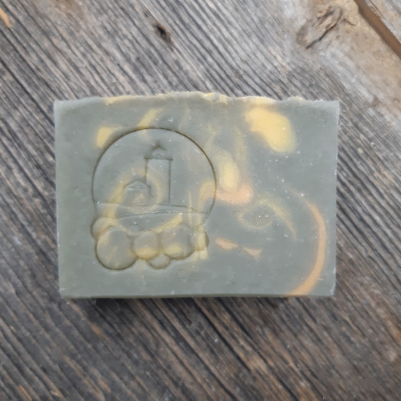 Puede incluir: Una pastilla de jab&oacute;n verde y amarilla con un logotipo de faro en relieve en la parte delantera. El jab&oacute;n est&aacute; sobre una superficie de madera.