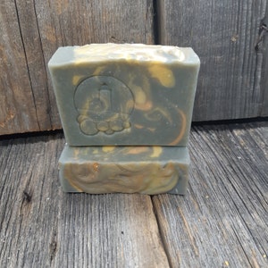 Puede incluir: Dos barras de jab&oacute;n hecho a mano con un patr&oacute;n de remolino en tonos de gris, amarillo y naranja. La barra superior tiene un logotipo circular con una letra estilizada "J" en su interior.