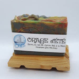 Puede incluir: Una pastilla de jab&oacute;n de leche de cabra verde, naranja y gris con una etiqueta blanca que dice "ORAGE d'&Eacute;T&Eacute; Savon au lait de ch&egrave;vre fait &agrave; la Main -Handmade Goat Milk Soap-". El jab&oacute;n est&aacute; sobre un soporte de madera.