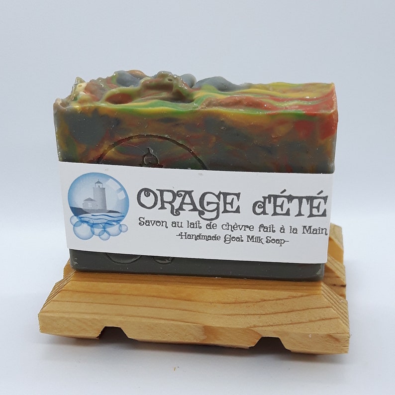Puede incluir: Una pastilla de jab&oacute;n de leche de cabra verde, naranja y gris con una etiqueta blanca que dice "ORAGE d'&Eacute;T&Eacute; Savon au lait de ch&egrave;vre fait &agrave; la Main -Handmade Goat Milk Soap-". El jab&oacute;n est&aacute; sobre un soporte de madera.