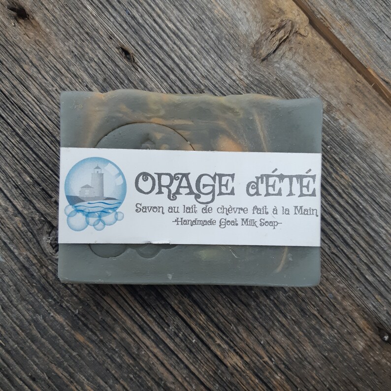Puede incluir: Una pastilla de jab&oacute;n de leche de cabra hecho a mano con una etiqueta que dice "ORAGE C'&Eacute;T&Eacute; Savon au lait de ch&egrave;vre fait &agrave; la Main -Handmade Goat Milk Soap-". El jab&oacute;n es de color verde oscuro con remolinos de naranja y marr&oacute;n.