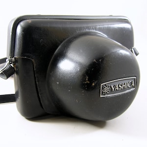 May include: Black leather camera case with the brand name "Yashica" in silver lettering. The case has a rounded shape and a shoulder strap. The case shows signs of wear and tear.