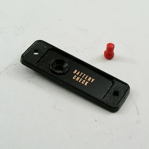 May include: A black rectangular component with the words "BATTERY CHECK" in gold lettering. A small red cylindrical piece sits nearby. The component has two holes and a circular indentation.