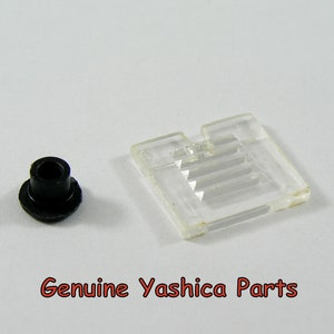 May include: A black plastic component and a clear plastic piece. The clear piece is rectangular with a series of parallel ridges. The text "Genuine Yashica Parts" is in red.
