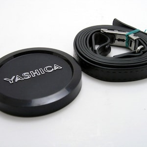 May include: A black camera lens cap with the word "YASHICA" printed in silver lettering, and a black camera strap with a silver buckle.