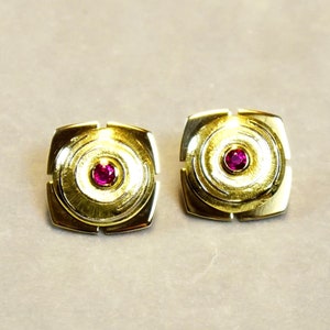 Könnte beinhalten: Ein Paar goldene Ohrringe mit quadratischem Design und einem kleinen roten Edelstein in der Mitte jedes Ohrrings.