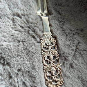 May include: Silver serving fork with two tines and an ornate handle. The handle features a detailed, symmetrical design with floral and fruit motifs. The fork is likely made of silver-plated metal and is designed for serving food.