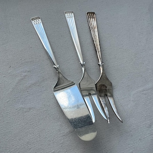 Op de afbeelding: Drie zilveren serveerbestekken: een taartschep en twee vorken. De taartschep heeft een plat, rechthoekig blad. De vorken hebben twee en drie tanden. Alle stukken hebben decoratieve handvatten met verticale lijnen.