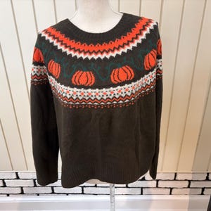 May include: A dark brown long-sleeve sweater featuring a crew neckline and a fair isle pattern across the chest. The design includes orange pumpkins with green vines, accented by white and orange geometric bands above and below the pumpkins.
