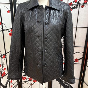 May include: A black quilted leather jacket with a collared neckline and button-up closure. The jacket features a diamond pattern and two front pockets. The jacket is displayed on a white mannequin.