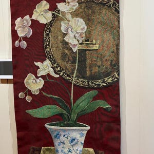 以下が含まれることがあります： 白い花と黄色の花、緑の葉を持つ鉢植えの蘭を描いたタペストリー。植物は青と白の模様の入った花瓶に入っており、濃い赤色の背景に配置されています。植物の上には、金のアクセントが付いた円形のデザインがあります。