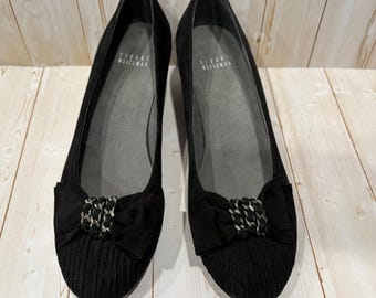 May include: A pair of black Stuart Weitzman flats. The shoes have a bow detail with a silver chain accent. The shoes have a textured design on the toe area.