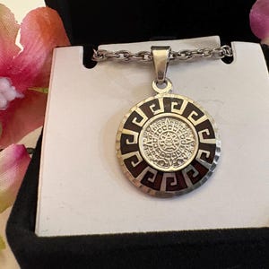 May include: A silver-toned circular pendant on a matching chain. The pendant features a central embossed Aztec calendar design surrounded by a dark brown border with a repeating silver Greek key pattern along the outer edge.