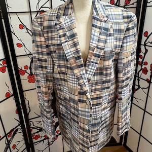 Puede incluir: Una blazer a cuadros en tonos azules, beige y blancos, exhibida en un maniquí. La chaqueta presenta un cierre clásico de dos botones y dos bolsillos delanteros. La tela tiene un diseño de patchwork.