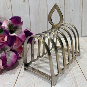 May include: A silver-colored, vintage toast rack with six arched slots. The rack has a decorative teardrop-shaped handle and is set against a light wood background. A bouquet of purple and pink flowers is to the left.