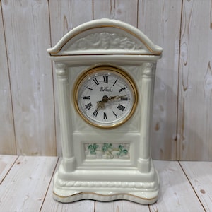 May include: Cream-colored ceramic mantel clock with a gold-tone clock face and trim. The clock face has black Roman numerals and the brand name "Bellack". The base features a shamrock design.