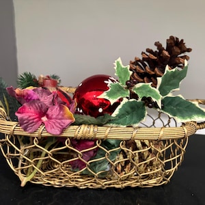 May include: A gold-toned wire basket with two handles, filled with a decorative arrangement featuring a shiny red ornament, a brown pinecone, green and white variegated holly leaves, and deep purple silk flower petals.
