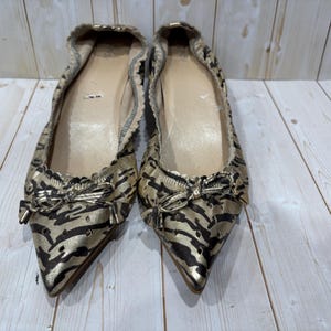 May include: A pair of gold and black patterned ballet flats with pointed toes and small bows. The shoes have a metallic sheen and a zebra-like pattern. The soles are a light tan color.
