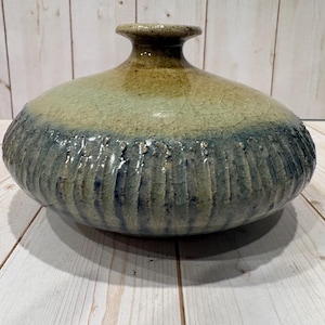 Puede incluir: Un jarrón de cerámica hecho a mano con un cuerpo bulboso y un cuello estrecho. El jarrón presenta un diseño texturizado y acanalado en la parte inferior, que transiciona a una superficie lisa y esmaltada. Los colores incluyen tonos de azul, verde y marrón.