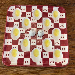Peut inclure: Un plat de service à carreaux rouges et blancs avec des supports à œufs farcis en céramique et deux petits salières et poivrières. Le plat est rectangulaire avec des coins arrondis. Les œufs farcis ont un jaune d'œuf jaune.