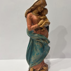 May include: A painted figurine of a figure with long brown hair wearing a white head covering, a salmon-colored long-sleeved garment, and a flowing blue draped cloth, holding a small child in their arms while standing on a hexagonal base.