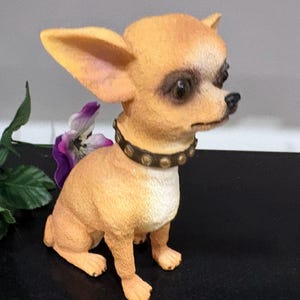 May include: A tan Chihuahua figurine with a studded collar. The dog has large ears and dark eyes. The figurine is sitting on a black surface, with a purple flower and green leaves in the background.