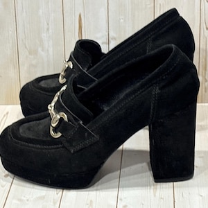 Puede incluir: Par de tacones altos negros con plataforma, tacón ancho y herrajes plateados. Los zapatos tienen un diseño tipo mocasín con punta redondeada y textura similar al ante. Los tacones miden aproximadamente 10 cm de alto.