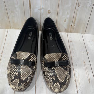 May include: A pair of Michael Michael Kors loafers. The shoes have a snakeskin pattern in shades of brown, tan, and black. Each shoe has a black bow with a silver-colored accent. The interior of the shoes is black.