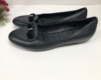 May include: A pair of black leather ballet flats featuring a rounded toe, a textured black fabric interior lining, and a black bow detail on the vamp accented with a small row of clear rhinestones. The shoes have a low profile and a flat sole.