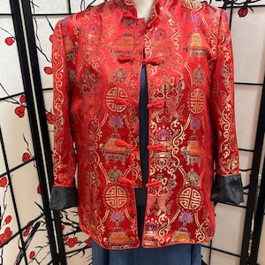 Puede incluir: Una chaqueta roja estampada con cuello mandarín y cierres de rana. La chaqueta presenta detalles dorados y morados, con puños negros. El diseño incluye motivos asiáticos tradicionales. La chaqueta está abierta, revelando una prenda azul debajo.