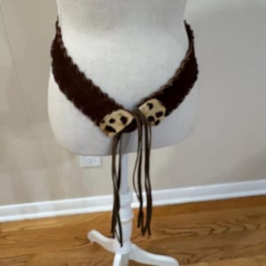 May include: A brown belt with a fringed design and leopard print accents. The belt is displayed on a mannequin and features long, dangling ties. The belt is made of a soft, textured material.