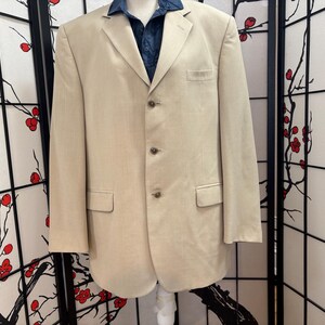 May include: A beige blazer with a notched lapel, three-button closure, and two front pockets. The blazer is paired with a dark blue collared shirt. The garment is displayed on a mannequin.