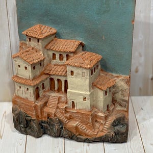 May include: A three-dimensional relief sculpture depicting a village scene. The buildings are painted in cream and brown tones, with terracotta-colored roofs. The background is a muted teal color, and the sculpture is mounted on a wooden base.