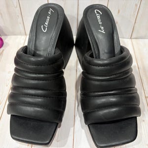 Pode incluir: Um par de sandálias pretas sem cadarços, com bico quadrado e uma tira grossa acolchoada. O nome da marca "Circus by Sam Edelman" está impresso em branco na palmilha. Os sapatos estão em uma superfície de madeira clara.