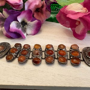 May include: A silver-toned metal bracelet featuring a series of connected links, each set with smooth, rounded, amber-colored stones. The two end links are larger and oval-shaped, while the central links are rectangular with rounded edges.