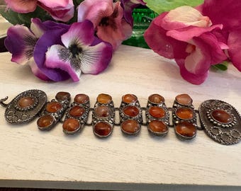 May include: A silver-toned metal bracelet featuring a series of connected links, each set with smooth, rounded, amber-colored stones. The two end links are larger and oval-shaped, while the central links are rectangular with rounded edges.