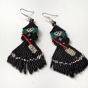 Made with black, red, silver, cream seed beads. The beads are hand sewn together to form a Halloween Spooky Witch with a broom. The witch has a pointed black hat and green hair. Her dress is black and silver. This picture is taken on a white surface.