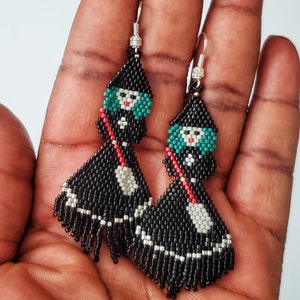 Made with black, red, silver, cream seed beads. The beads are hand sewn together to form a Halloween Spooky Witch with a broom. The witch has a pointed black hat and green hair. Her dress is black and silver.