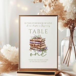 May include: A gold-framed table number card with the text "IN THIS STORYBOOK OF LOVE" and "TABLE ONE." The card features a watercolor illustration of stacked books tied with a ribbon and floral accents. The card is displayed on a gold stand.