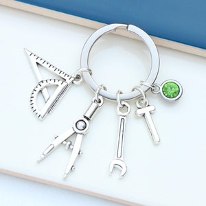 Könnte beinhalten: Silberner Schlüsselanhänger mit Kompass, Winkelmesser, Schraubenschlüssel und Buchstaben T-Anhängern. Ein grüner Edelstein ist am Ring befestigt.