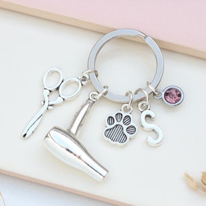以下が含まれることがあります： 足跡のチャーム、Sの文字のチャーム、はさみのチャーム、ヘアドライヤーのチャーム、ピンクのラインストーンのチャームが付いたシルバーのキーホルダーです。
