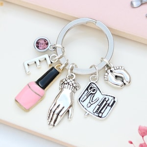 Può includere: Portachiavi color argento con ciondoli relativi alla cura delle unghie. I ciondoli includono una bottiglia di smalto rosa, una mano, forbici, un libro e impronte di piedi di bambino. È attaccata anche una lettera "E" e una gemma rosa.