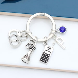 May include: Silver keyring with a variety of charms, including handcuffs, a torch, a phone, a letter 'P', and a blue rhinestone.